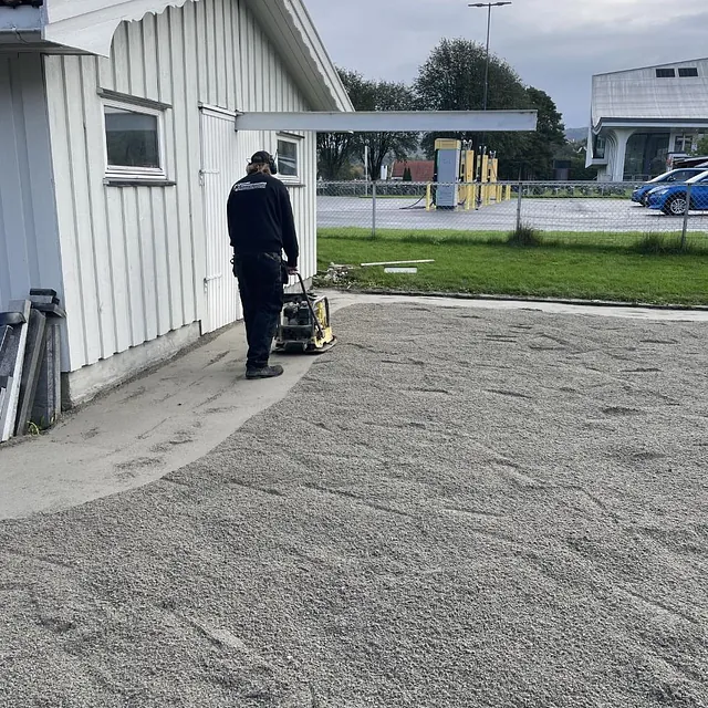 Person som arbeider på en gruslagt gårdsplass foran et hvitt hus.