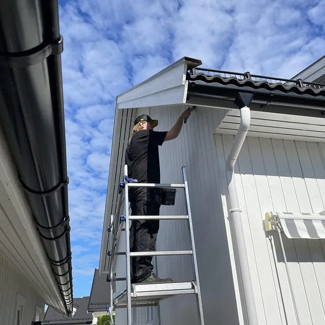Person står på stige og arbeider ved takutstikk på hvitt hus under blå himmel.
