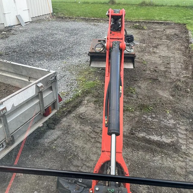 Liten rød graver eller minigraver i arbeid med å grave opp et jordområde ved en garasje.