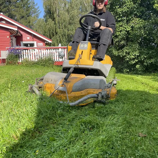 Person kjører sitteklipper og klipper høyt gress på grønt uteområde ved bolig.